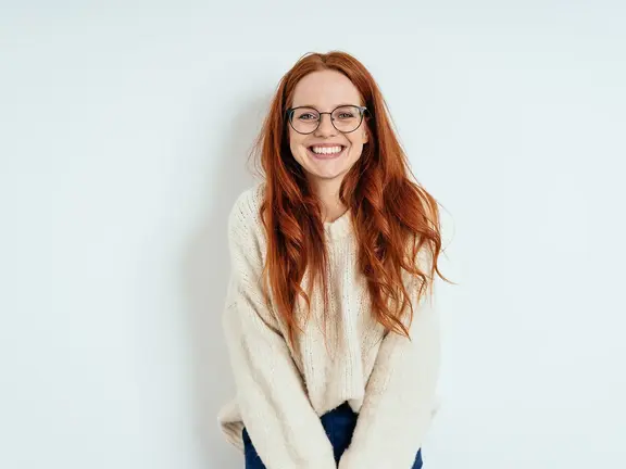 Lächelnde Frau mit langen roten Haaren und Brille, die einen cremefarbenen Pullover trägt und vor einem schlichten weißen Hintergrund steht.
